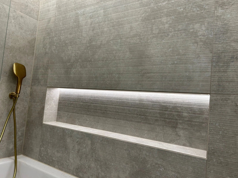 Gray tiled shower wall with a gold shower head and recessed lighting.