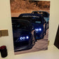 A vertical led photographic print of three dark sports cars lined up on a dirt road, displayed leaning against a light-colored wall on a white surface.