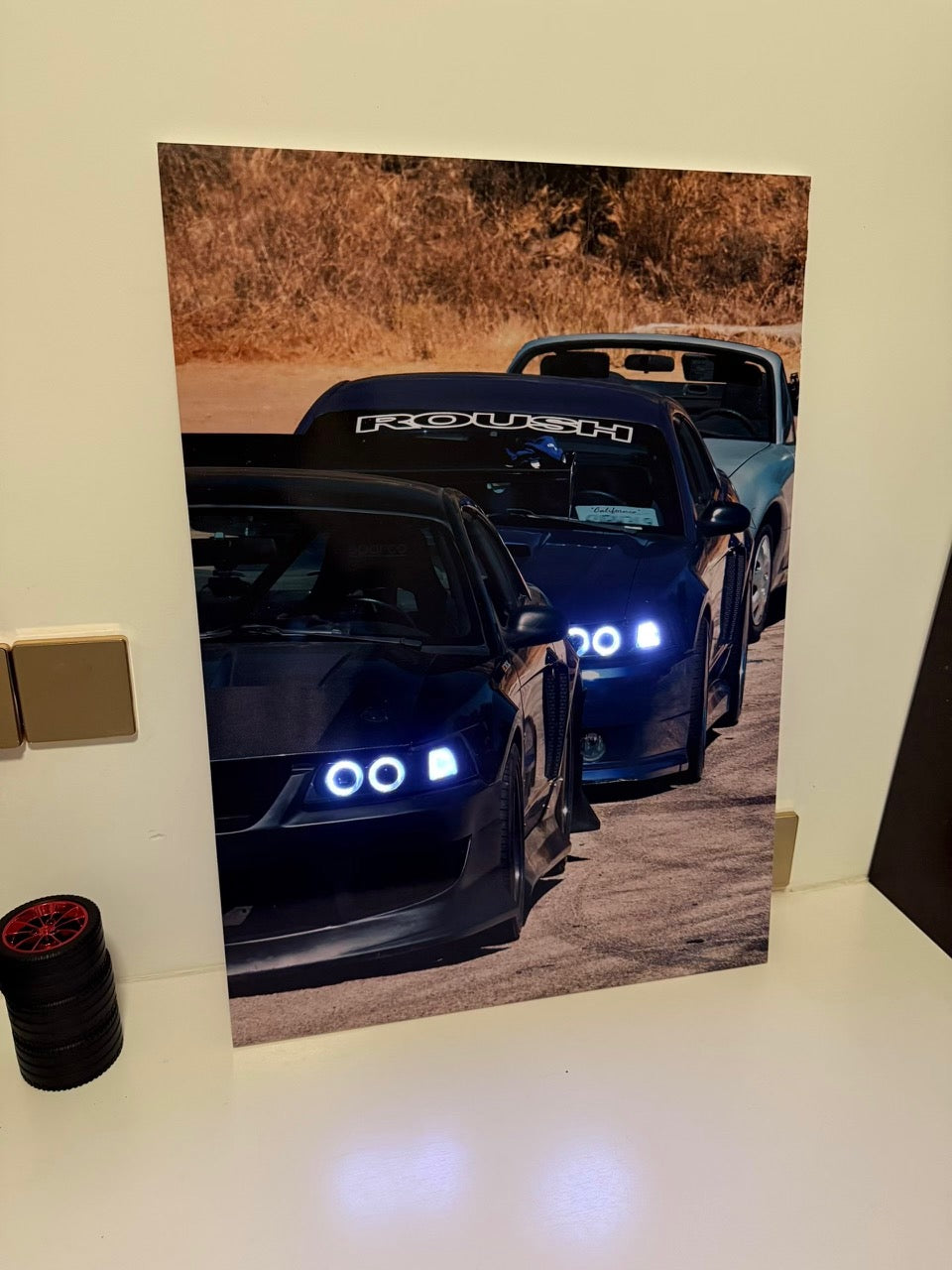 A vertical led photographic print of three dark sports cars lined up on a dirt road, displayed leaning against a light-colored wall on a white surface.
