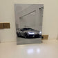 A vertical rectangular wall art print or poster leaning against a white wall on a white surface, showing a black-and-white photo of a silver sports car in a minimalist interior. No person or animal is present.