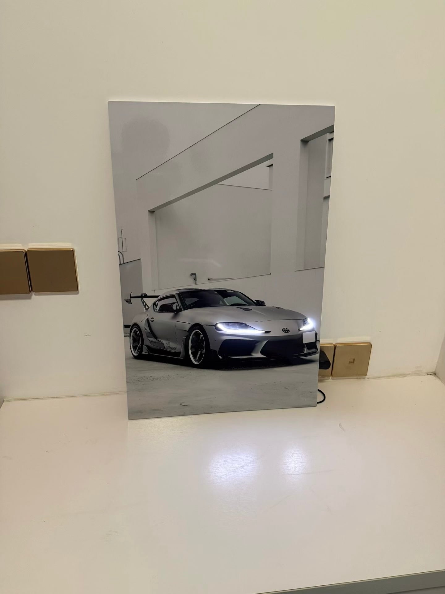 A vertical rectangular wall art print or poster leaning against a white wall on a white surface, showing a black-and-white photo of a silver sports car in a minimalist interior. No person or animal is present.