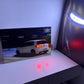 A rectangular wall-mounted display or screen showing a white car at night, installed on a white desk against a blue-lit wall. A large dark poster or panel with a bright horizontal light is visible on the right.