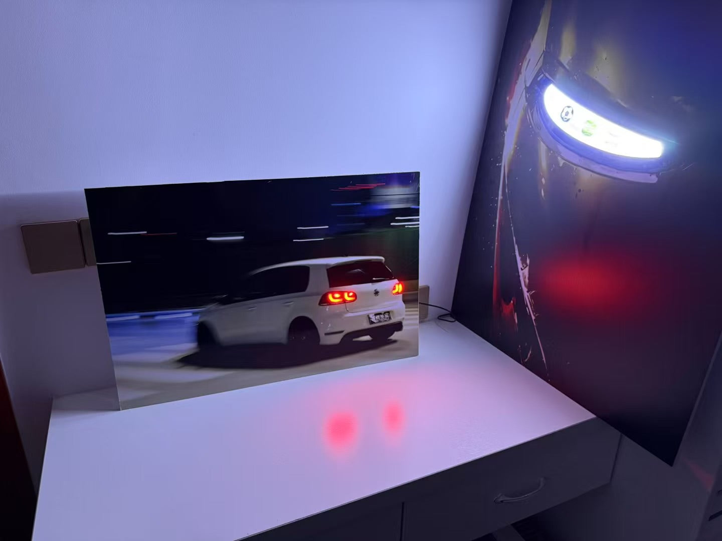 A rectangular wall-mounted display or screen showing a white car at night, installed on a white desk against a blue-lit wall. A large dark poster or panel with a bright horizontal light is visible on the right.