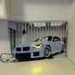 A rectangular wall-mounted light or lamp featuring a printed image of a light blue sports car in a garage, shown turned on and casting a warm yellow glow on the white surface below it.