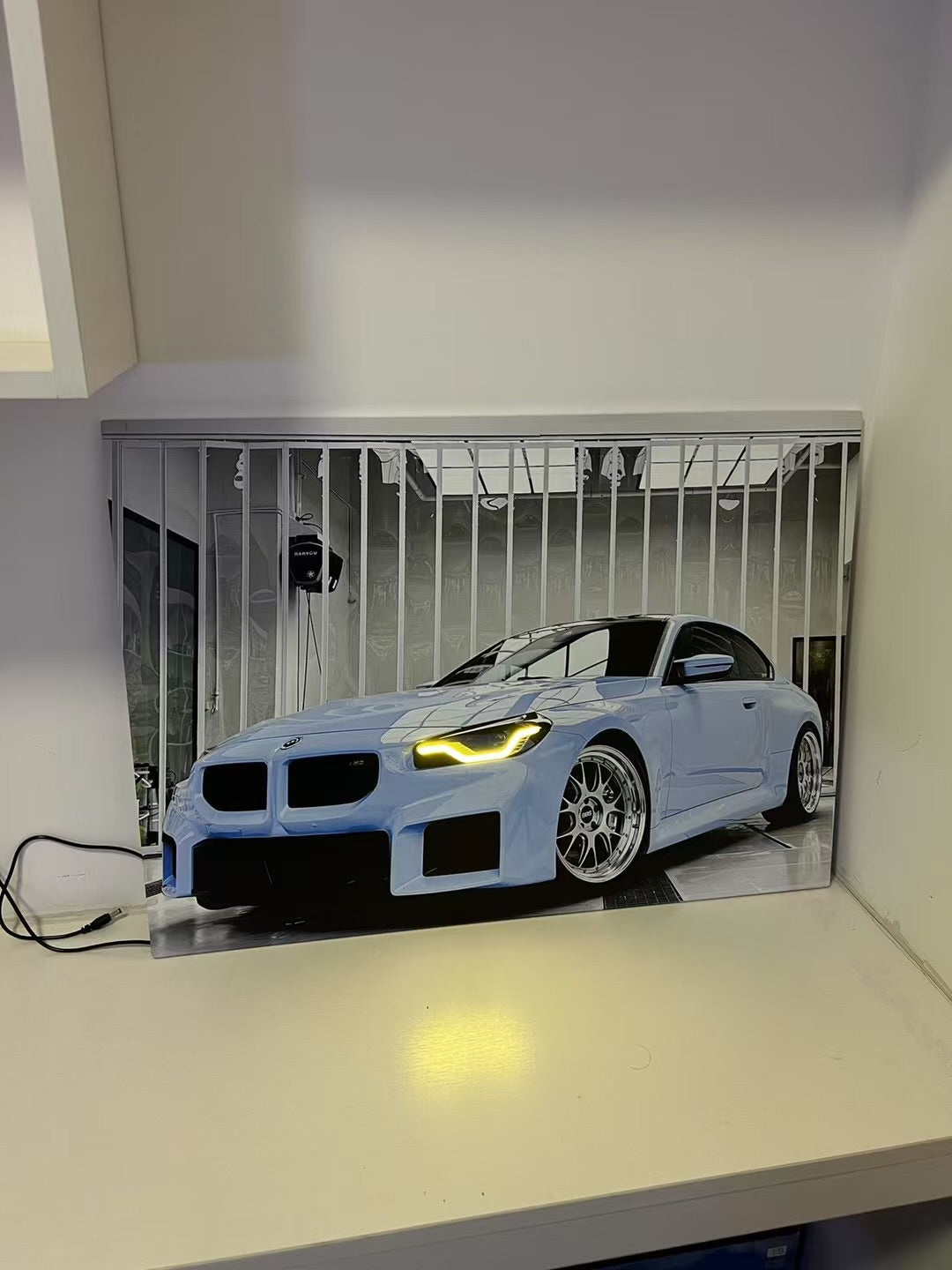 A rectangular wall-mounted light or lamp featuring a printed image of a light blue sports car in a garage, shown turned on and casting a warm yellow glow on the white surface below it.