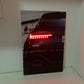 A vertical wall art print or poster leaning against a white wall on a white shelf, showing the rear of a black car with illuminated red taillights.
