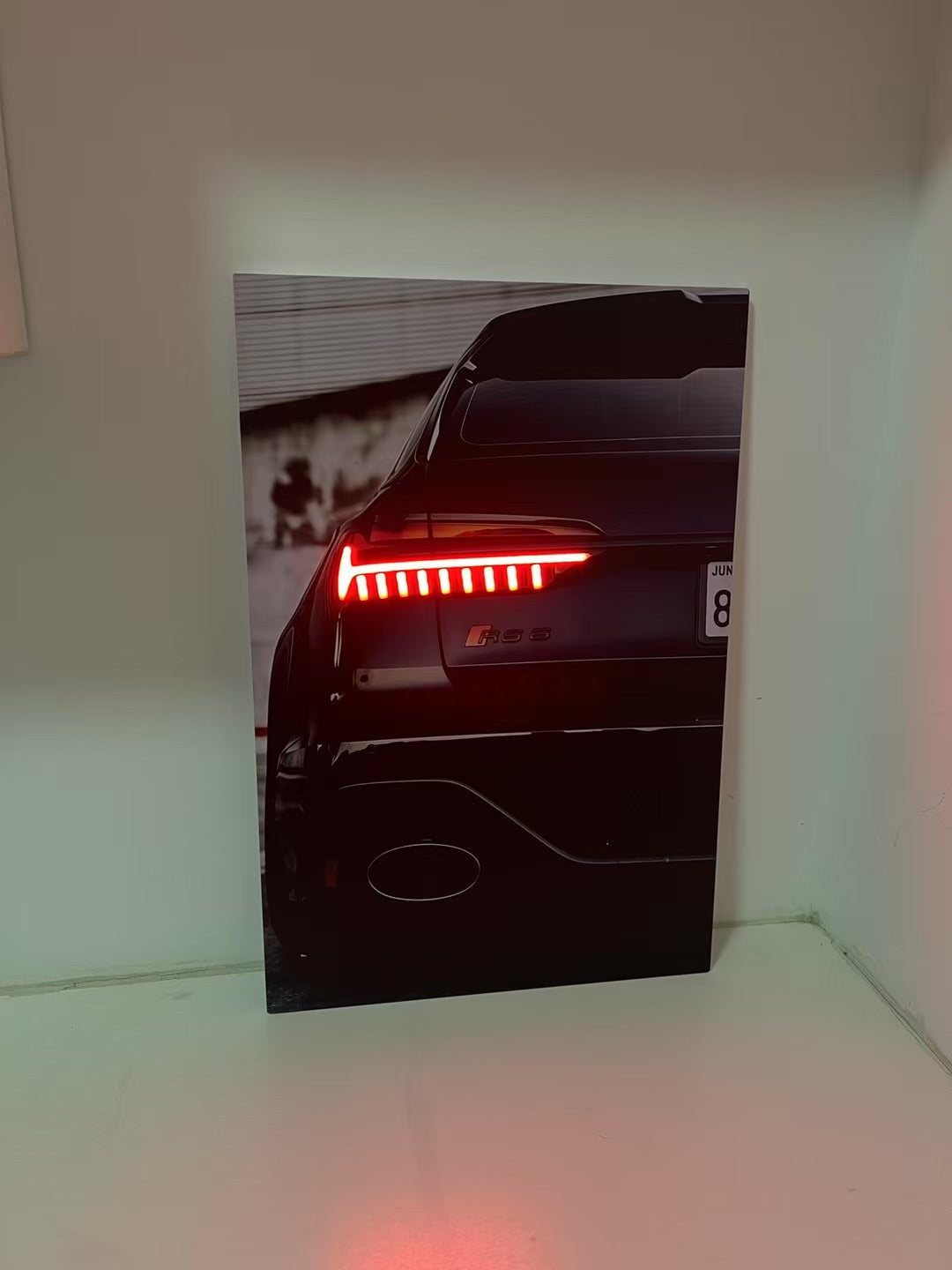A vertical wall art print or poster leaning against a white wall on a white shelf, showing the rear of a black car with illuminated red taillights.