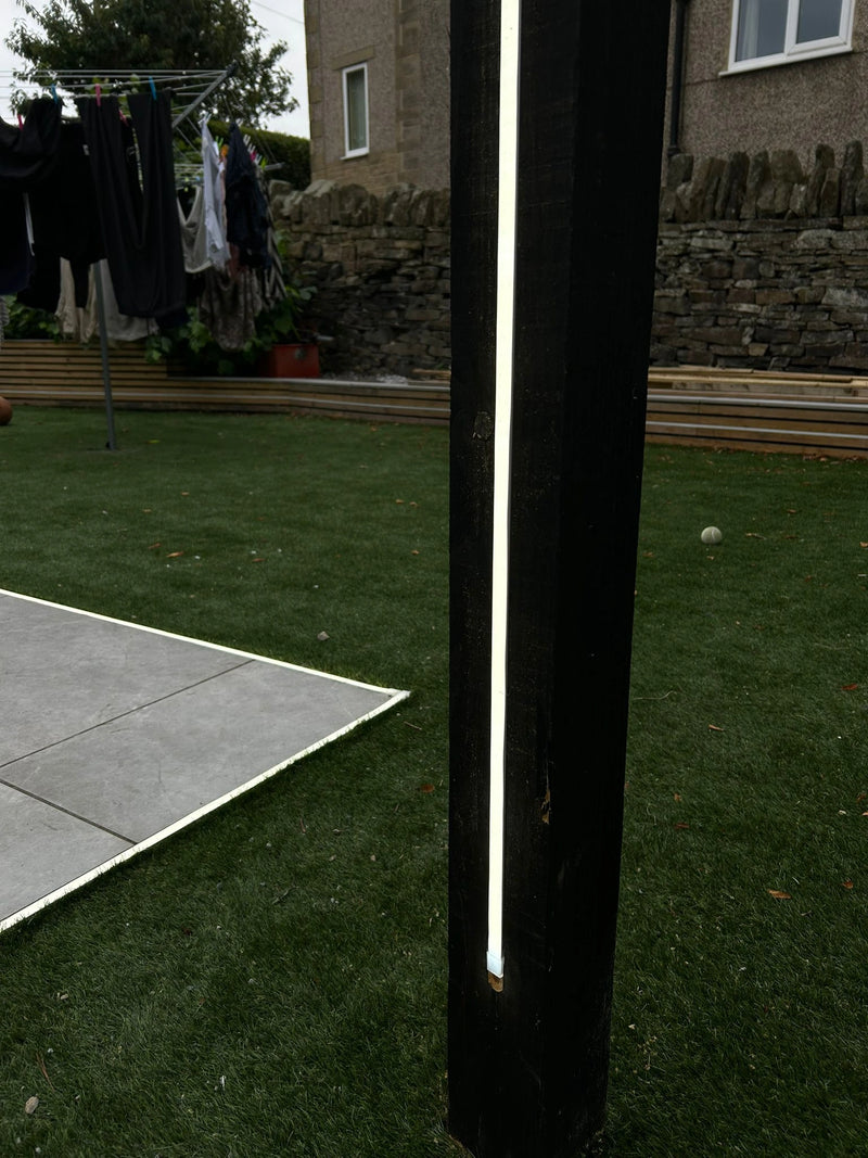 Neon light fixture on a wooden post in a backyard setting with a stone wall and clothesline in the background.