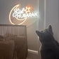 Grey cat looking at a neon 'Eid Mubarak' sign on a wall.
